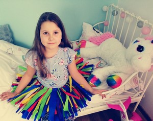 Dziewczyna siedząca na łóżku w spódniczce wykonanej z kolorowych pasków bibuły.