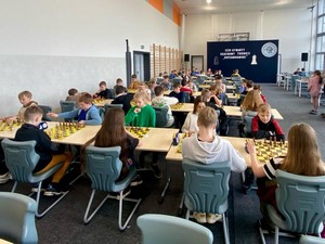 Uczestnicy XXVI Otwartego Szachowego Turnieju Kopernikowskiego siedzą przy stolikach i grają w szachy na sali gimnastycznej.
