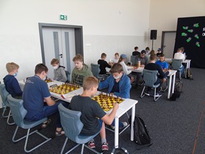 Uczestnicy turnieju szachowego siedzą przy stolikach (planszach szachowych) na sali gimnastycznej.