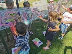 Dzieci z oddziałów przedszkolnych malują farbami obrazki na folii rozciągniętej pomiędzy drzewami.