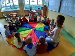 Dzieci z wychowawczynią siedzą w sali na podłodze wokół chusty animacyjnej.
