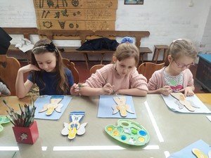 Dzieci z klasy 1b siedzą przy stole i malują wzory kaszubskie na drewnianych deseczkach.