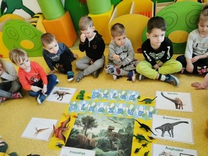 Dzieci z oddziału przedszkolnego (3 - latki) siedzą na podłodze wraz z wychowawczynią, która przedstawia im materiały związane z dinozaurami.