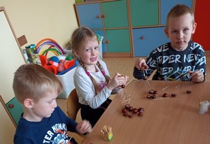 Dzieci siedzą przy stoliku i tworzą kompozycje z kasztanów i wykałaczek.