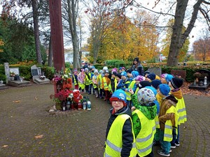 Dzieci z oddziałów przedszkolnych stoją przy krzyżu na cmentarzu.