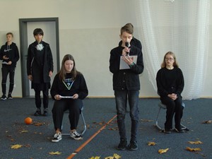 Uczniowie klas 8 na sali gimnastycznej recytują poezję.