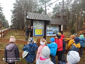 Uczniowie z klasy 1a przy tablicy informacyjnej słuchają informacji na temat zachowania w lesie.