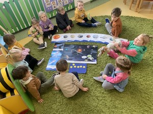 Dzieci z oddziału przedszkolnego (3 latki) siedzą na podłodze przed planszami edukacyjnymi przedstawiającymi planety.