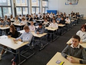 Uczniowie z klas 8 siedzą przy stolikach na sali gimnastycznej przed standaryzacją arkusza egzaminacyjnego z matematyki.