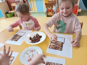 Dzieci z oddziału przedszkolnego (4 - latki) siedzą przy stoliku i wykonują pracę plastyczną za pomocą czekolady.
