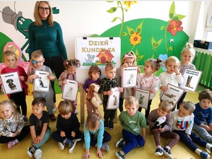 Dzieci z oddziałów przedszkolnych (3 latki) wraz z wychowawczynią prezentują się do zdjęcia na tle plakatu "Dzień Kundelka".