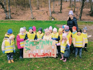 Dzieci z oddziału przedszkolnego (4 – latki) stoją wraz z wychowawczynią w Gaju Świętopełka i prezetnują się do zdjęcia prezentując plakat "Sprzątamy dla Polski".